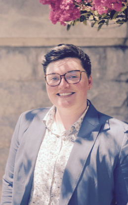 Photo of person with glasses in a sunlit area with a flowering tree. They are wearing a suit jacket and buttoned shirt and smiling.