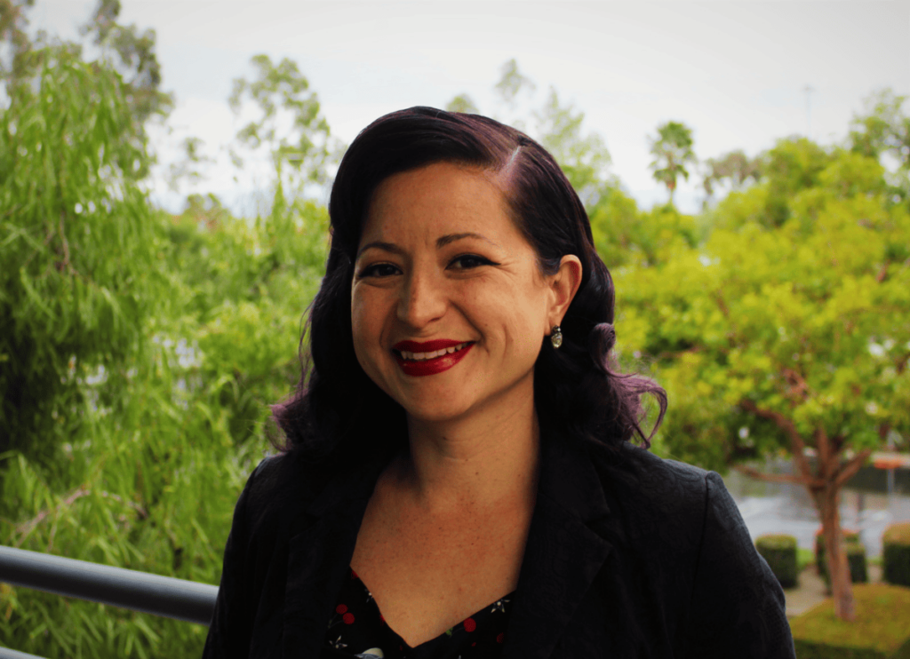 photo of woman with long curled hair and bright red lipstick smiling outside in front of background of trees